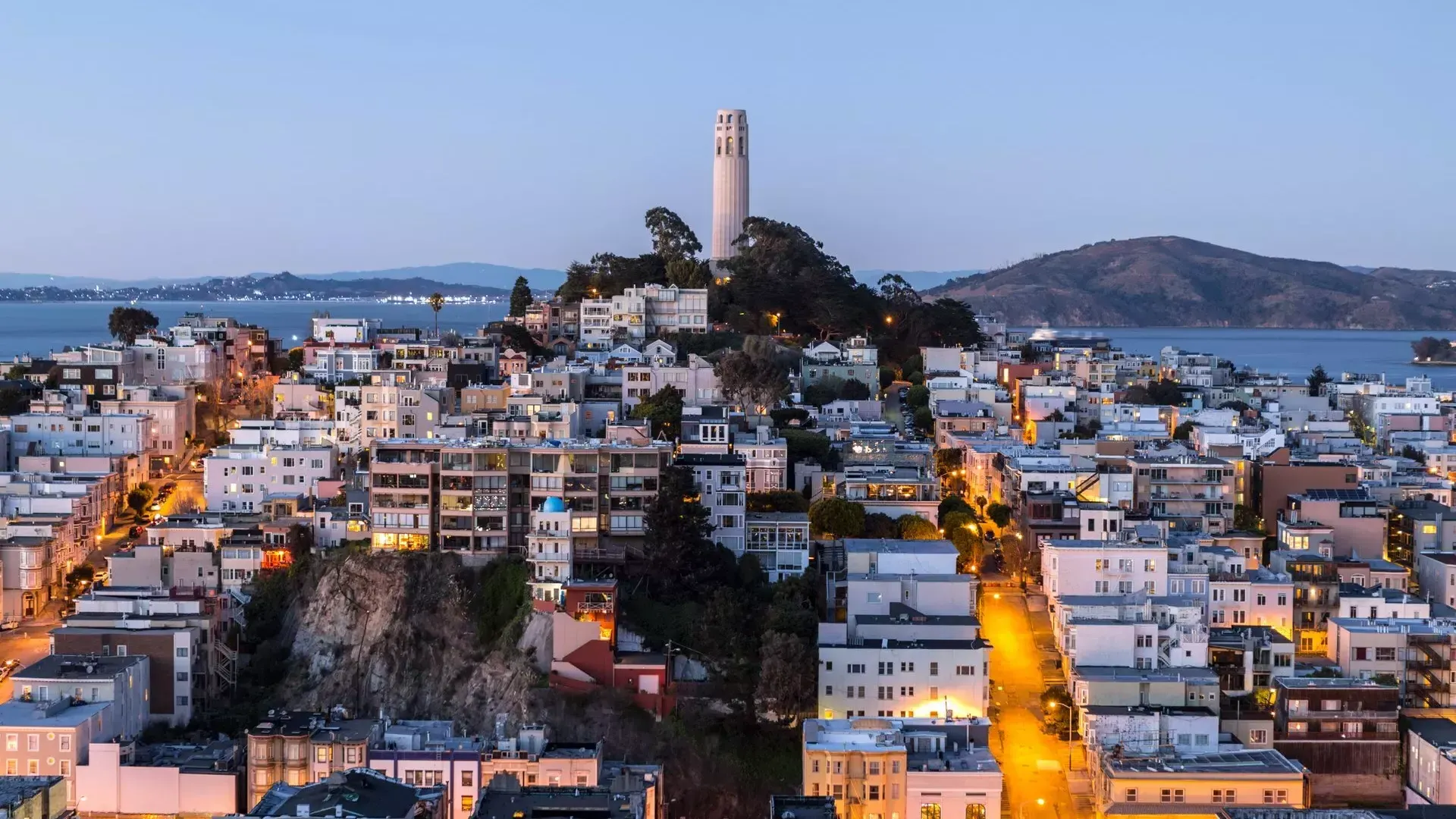 夕暮れ時のサンフランシスコのCoit Tower 。その前には明かりの灯った通り、背後にはサンフランシスコ湾が広がっています。